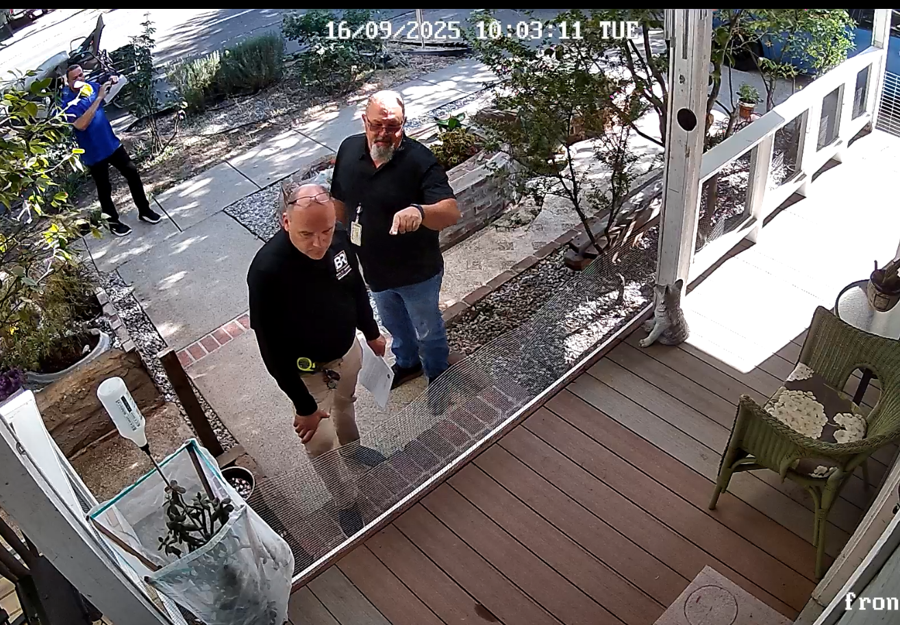 Wide front-door CCTV screenshot at 10:03:11 showing blue-shirt official near the sidewalk while other officials approach the porch.